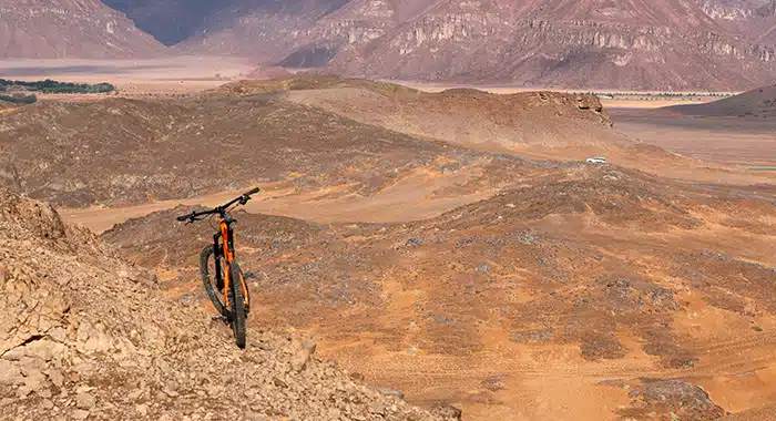 Cyclist riding through the desert trail at Alula Bike Park, with dramatic rock formations and vast landscapes in the background.