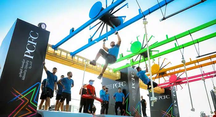 Participants navigating a high-ground wheel obstacle by pulling and swinging through during the Government Games at Kite Beach, Dubai, showcasing agility and strength.