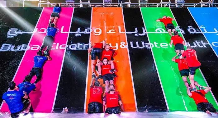 Team members scaling a high wall together during the Government Games at Kite Beach, Dubai, demonstrating teamwork and physical strength.