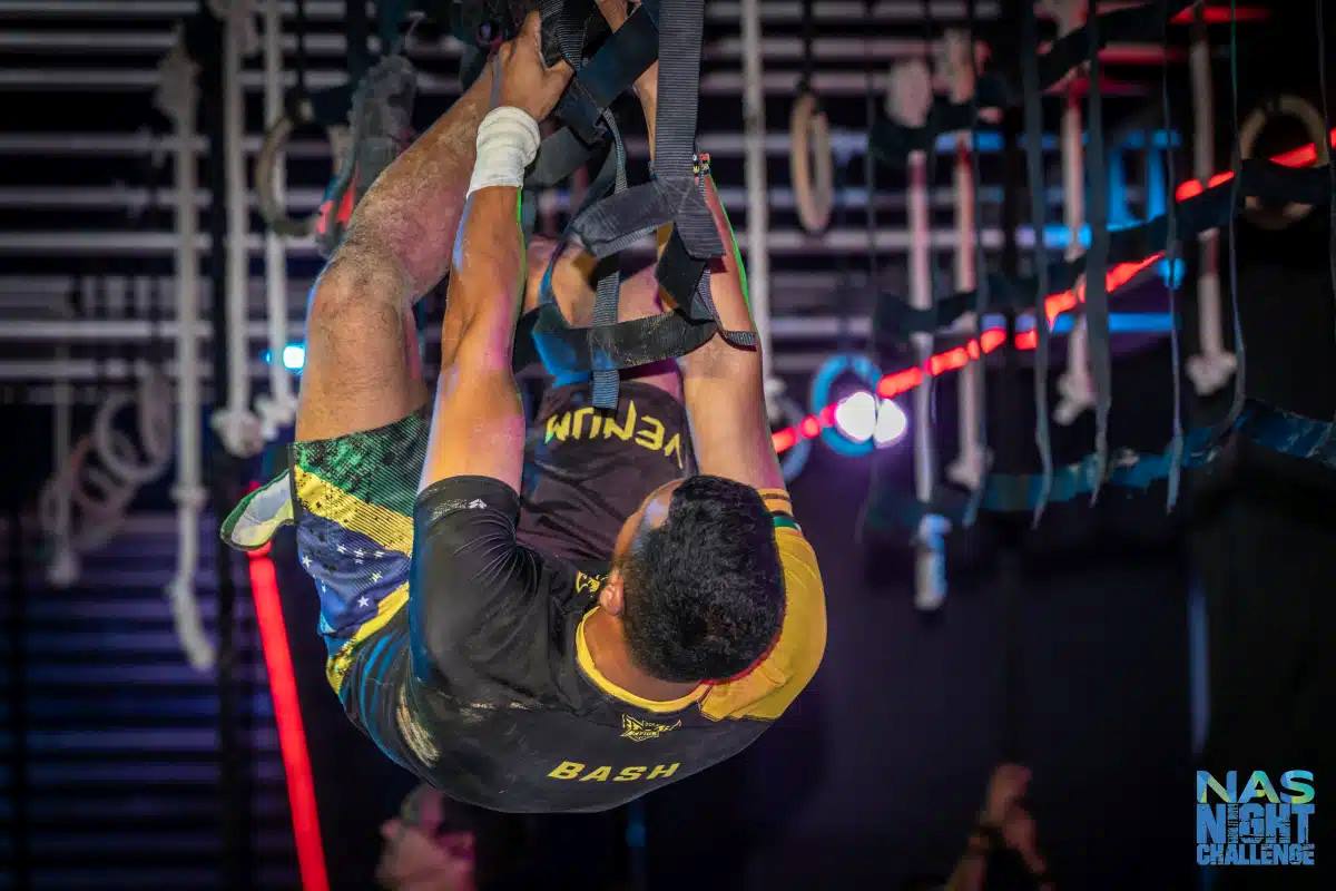 Participant scaling a challenging rope obstacle at the NAS Night Challenge event, featuring vibrant lighting and intense athletic focus.