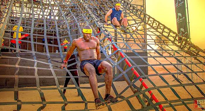 Competitor climbing a wall rope obstacle during the Desert Warrior Challenge, surrounded by the desert's sandy terrain.