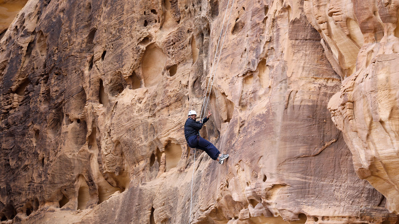 Adventurer abseiling down a towering mountain cliff in AlUla, Saudi Arabia, enjoying stunning desert views.